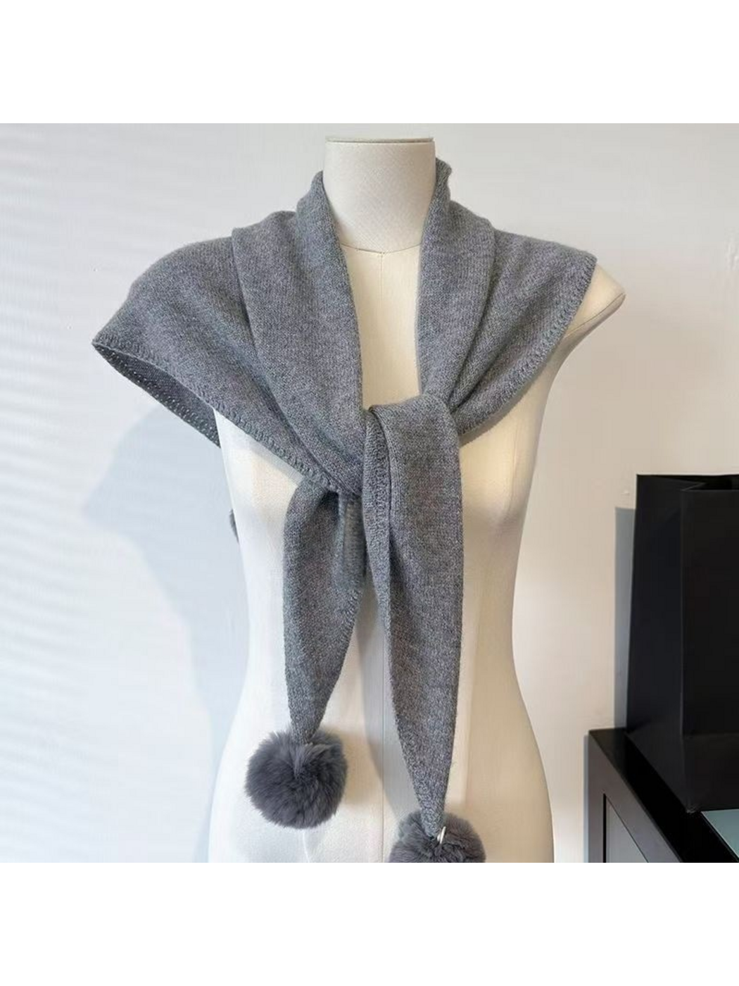 Gray knit scarf draped across a mannequin's shoulders with unequal hanging ends and a partly cropped pom-pom