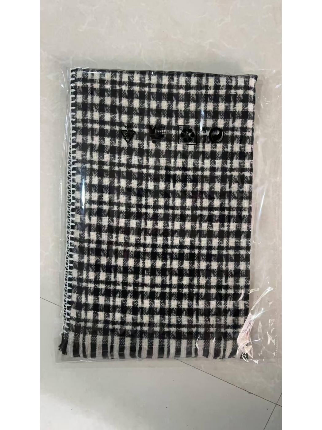 Black plaid scarf folded inside clear packaging, with white edge stitching and fringe visible