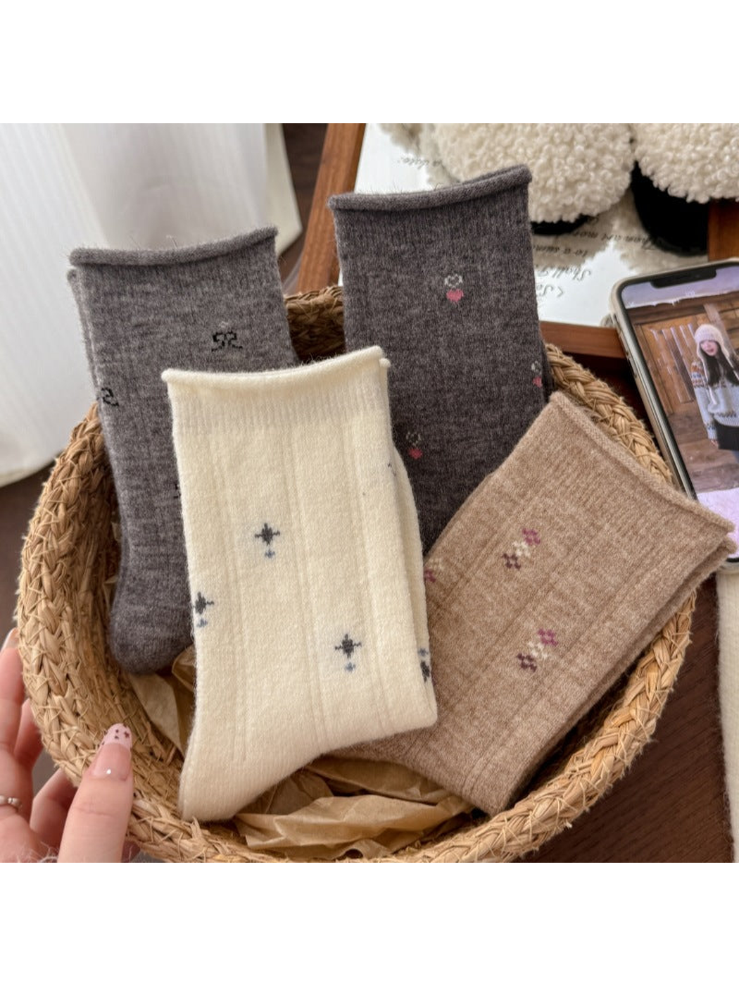 Four folded patterned socks arranged in a woven basket beside a phone displaying a person.