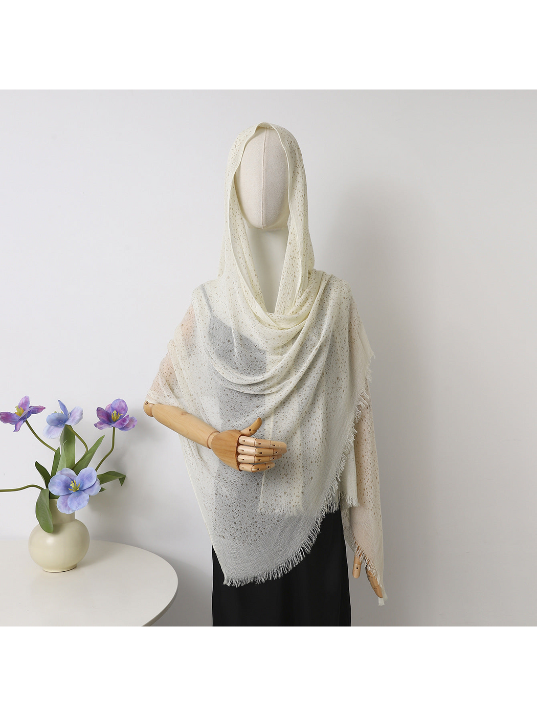 Ivory openwork scarf with fringed edges draped over a mannequin beside purple flowers