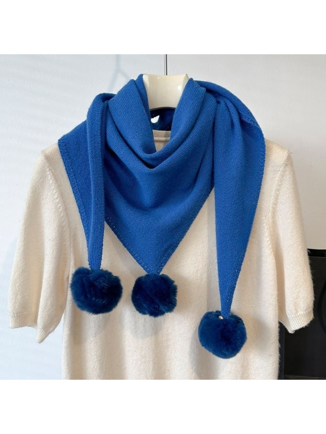 Blue knit triangle scarf looped over a cream top, with three pom-poms visible