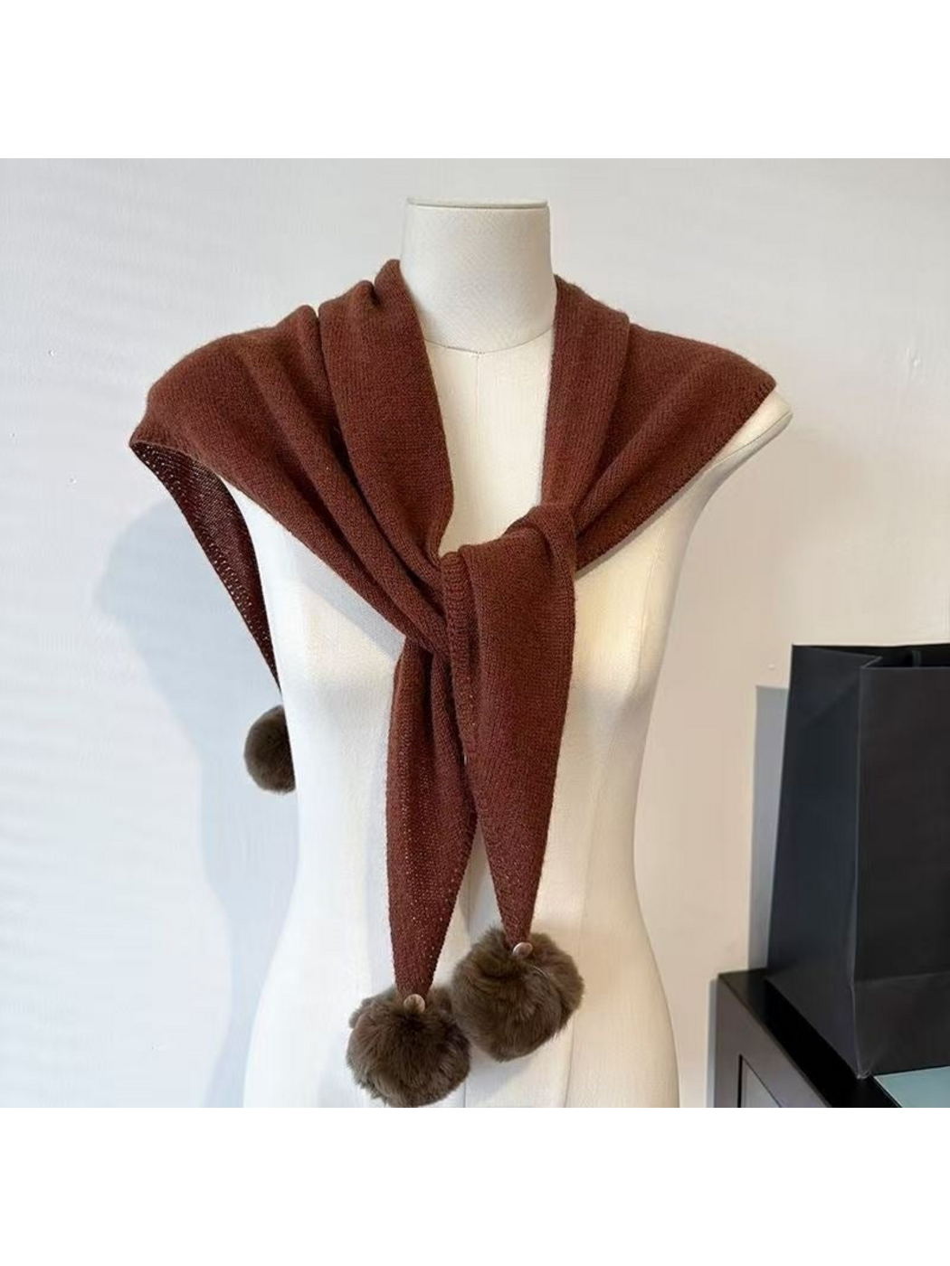 Brown knit scarf loosely tied across a mannequin's shoulders, with pom-pom ends