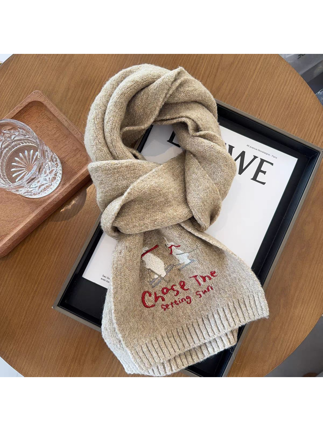 Beige scarf looped on a tabletop, showing skiing figures, lettering and ribbed ends