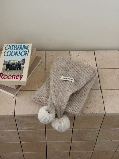 Beige knit scarf folded with two off-white pom-poms and an attached label
