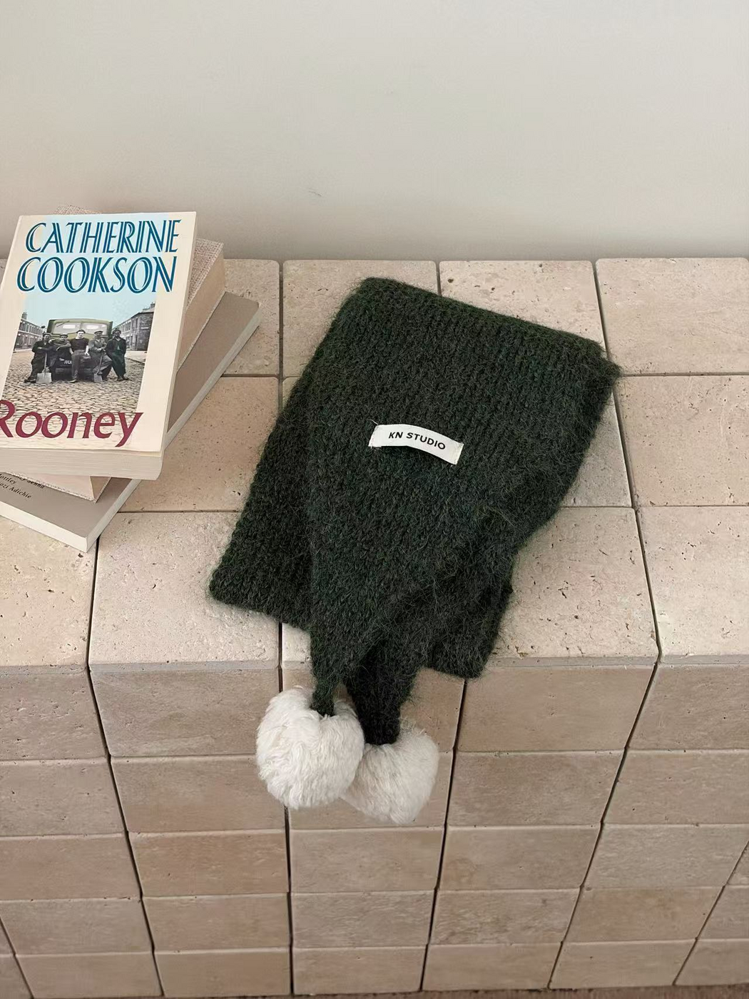 Green knit scarf folded with two white pom-poms and an attached label