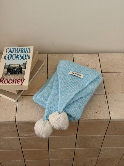 Blue knit scarf folded with two off-white pom-poms and an attached label