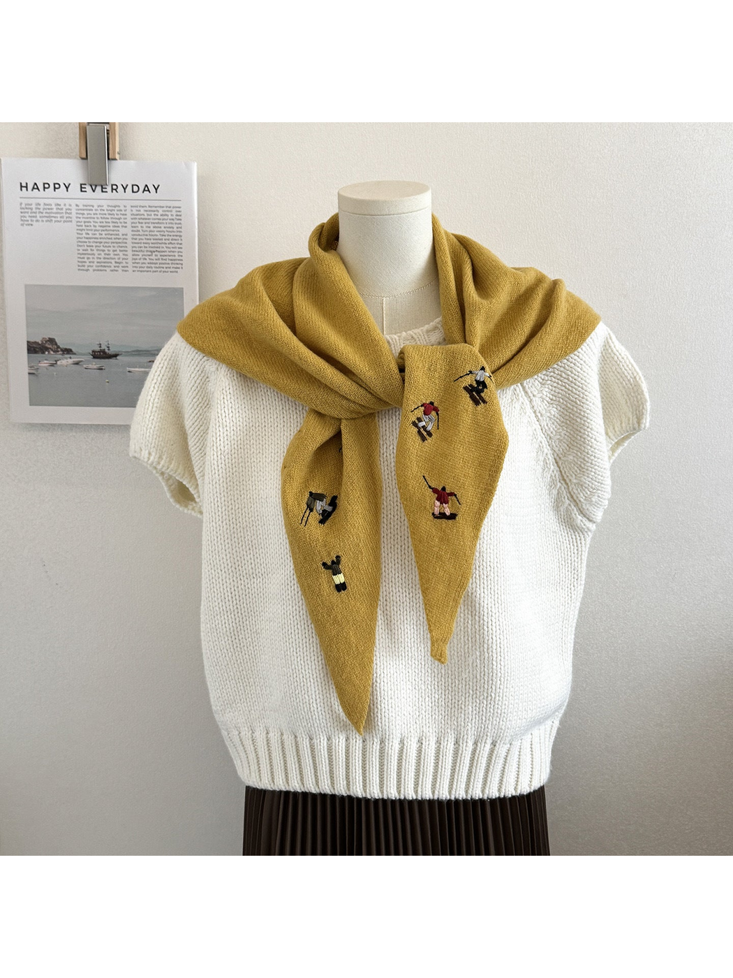 Mustard knit scarf with embroidered winter-sport figures, tied over a white top