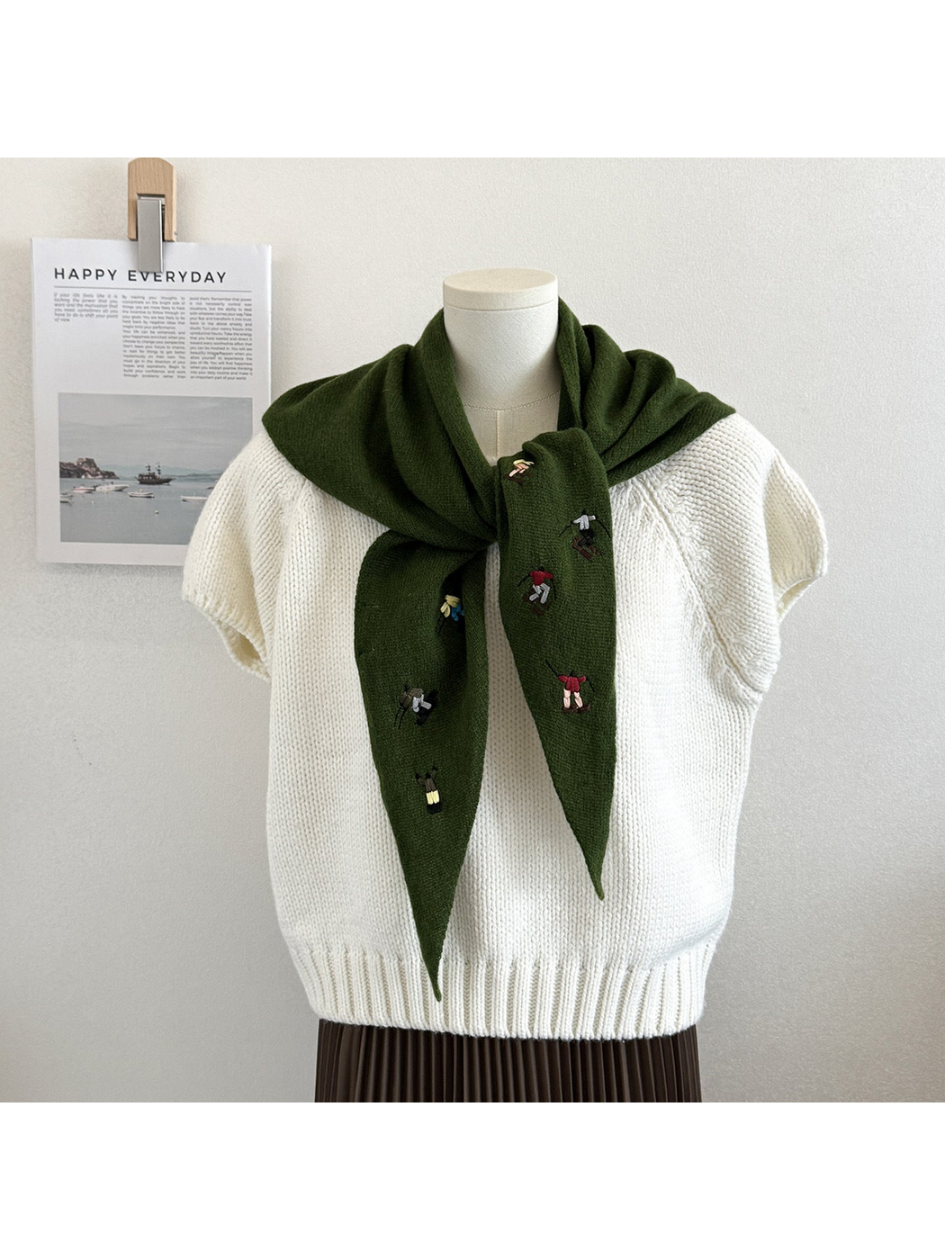 Green knit scarf with embroidered winter-sport figures, tied over a white top