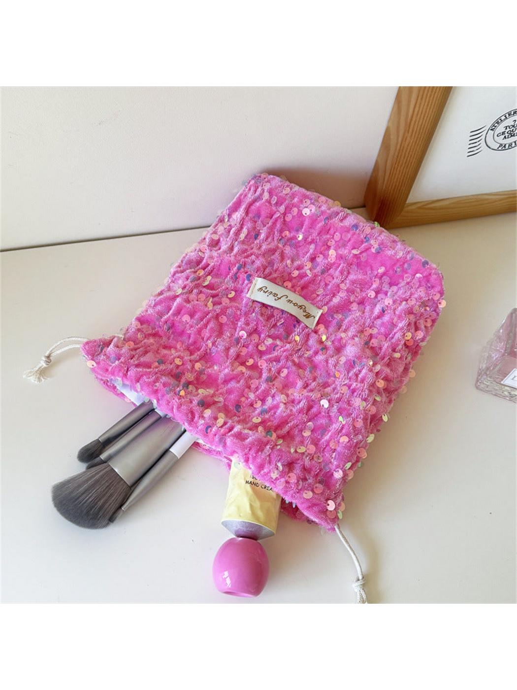 Pink sequined pouch spread open with makeup brushes and a cream tube extending from the opening