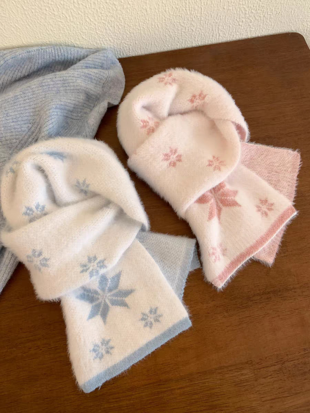 White scarves with blue and pink snowflake motifs displayed together on a wooden table