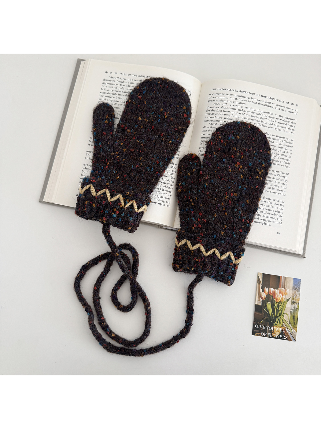 Flecked mittens laid across an open book, with inward-facing thumbs and a looped connecting cord