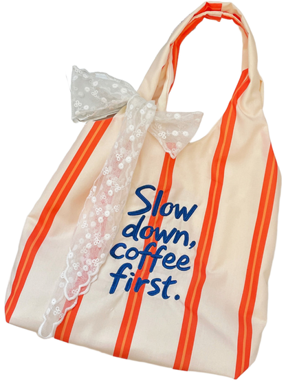 Orange tote bag with narrow orange stripes and blue slogan lettering, displayed with a white lace bow