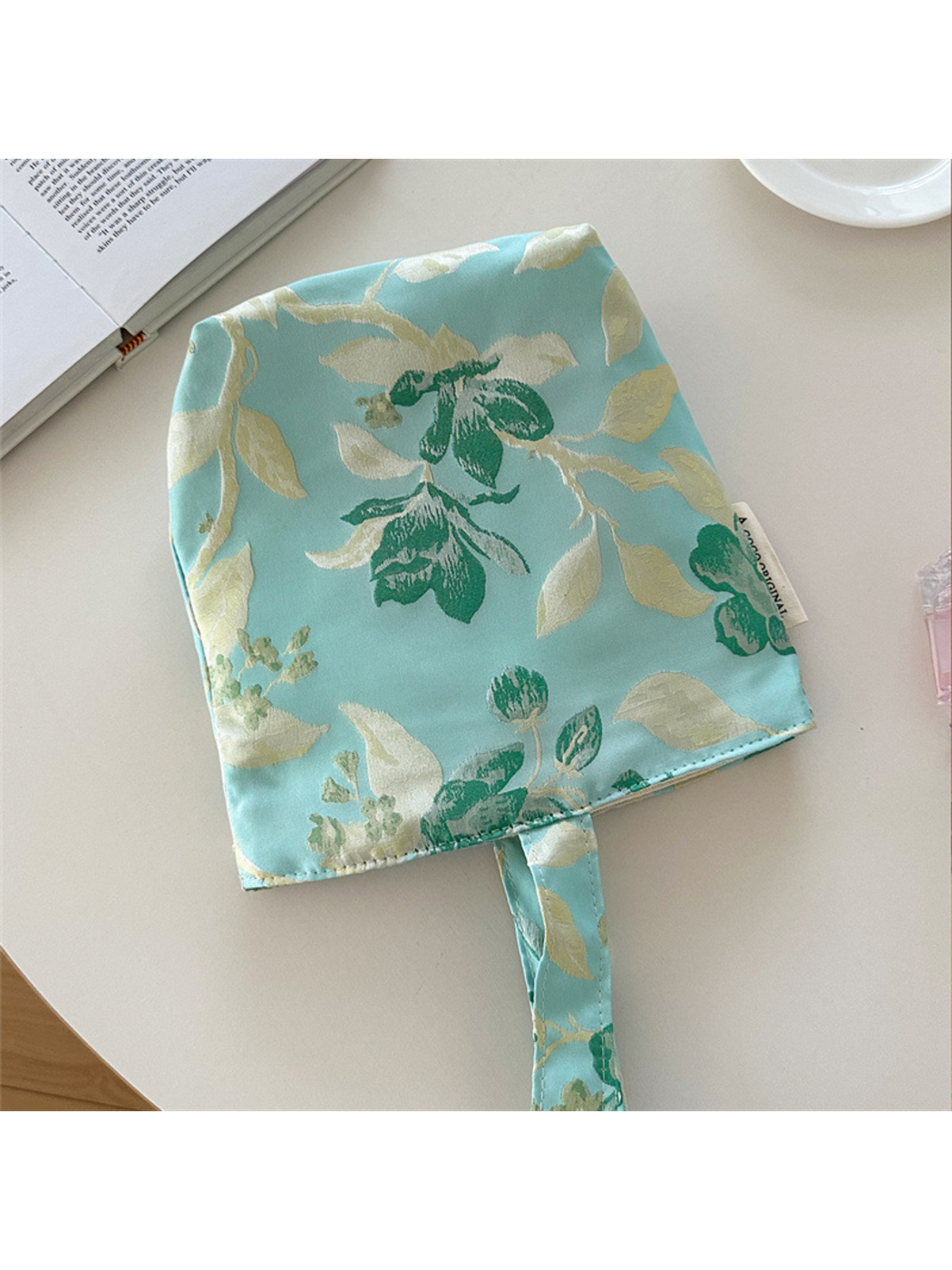 Turquoise bag laid flat, showing green flowers and cream branches with the handle partly outside the frame