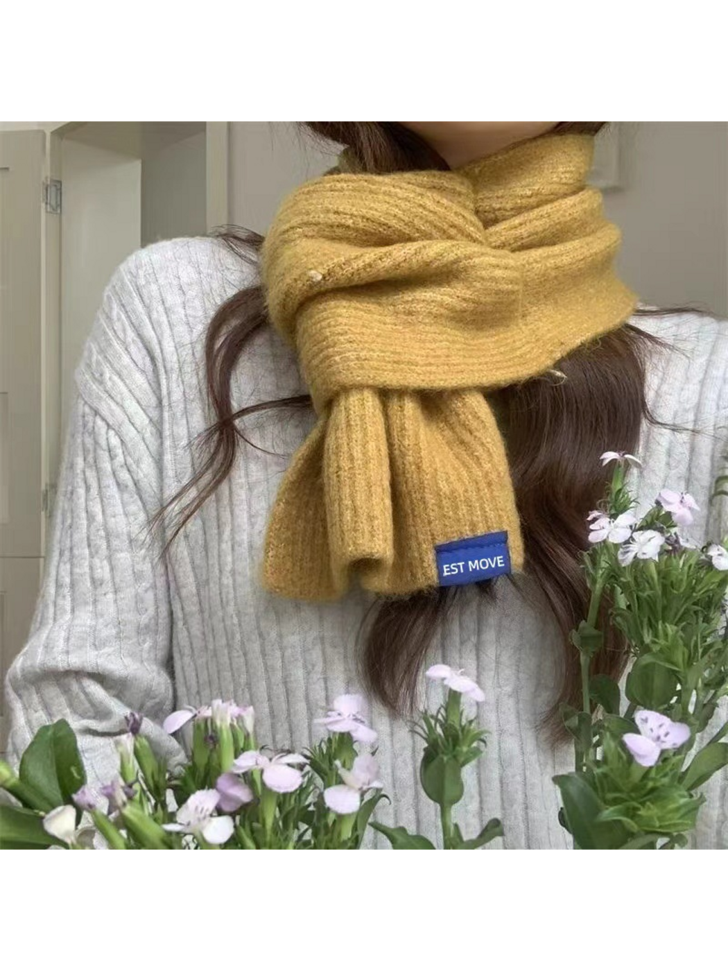 Yellow ribbed scarf worn around the neck above a gray sweater, with flowers in the foreground