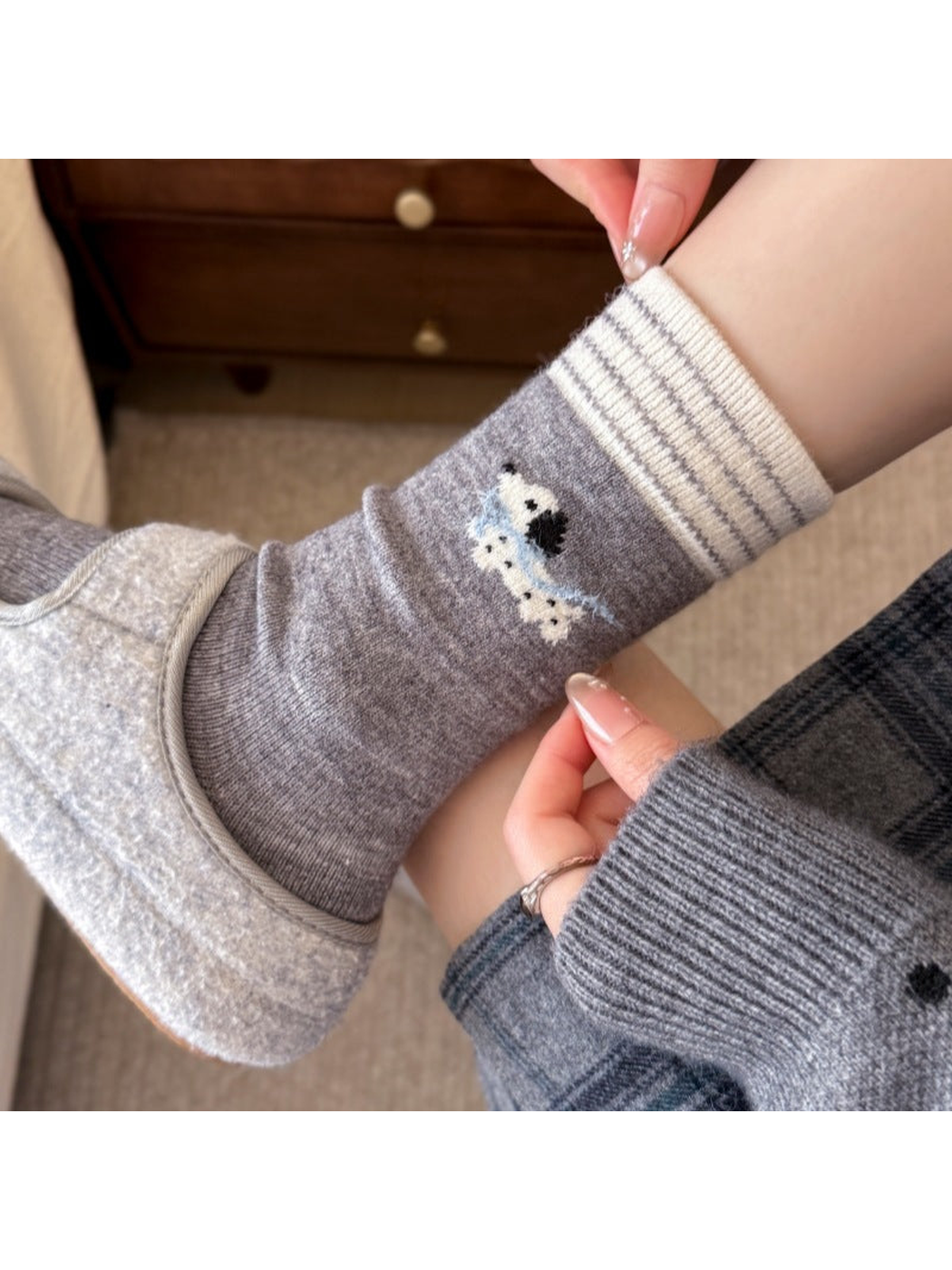 Light gray sock with a striped cream cuff and a dog with a blue scarf, worn with a slipper while hands pull the cuff and fabric.