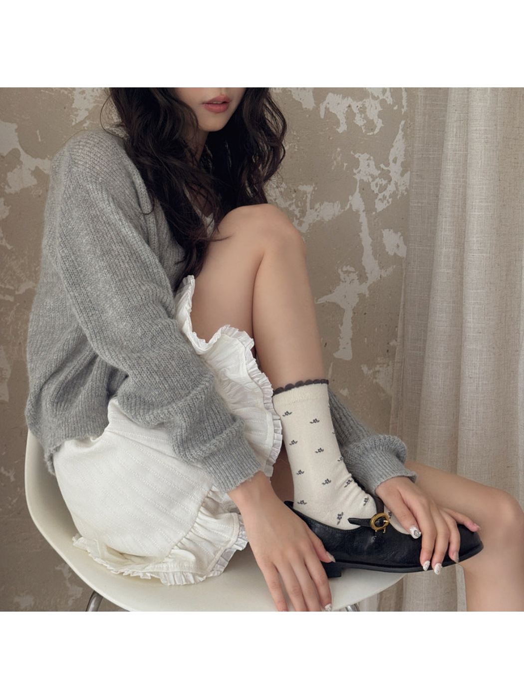 Seated model wearing an ivory floral sock with a contrasting scalloped cuff and a black buckle shoe.