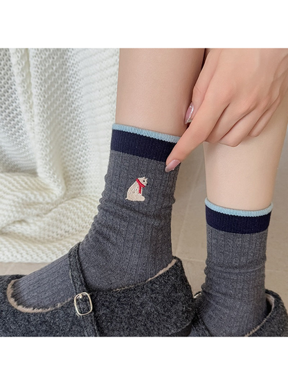 Dark gray Bear socks worn with dark slippers, with a finger pointing beside the embroidered motif.