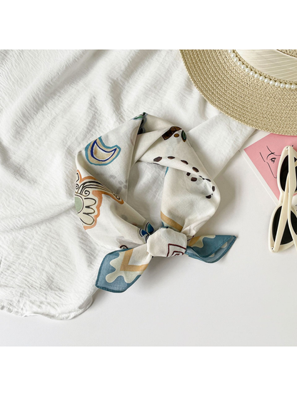 Abstract-print square scarf tied in a loose knot beside a sun hat and sunglasses