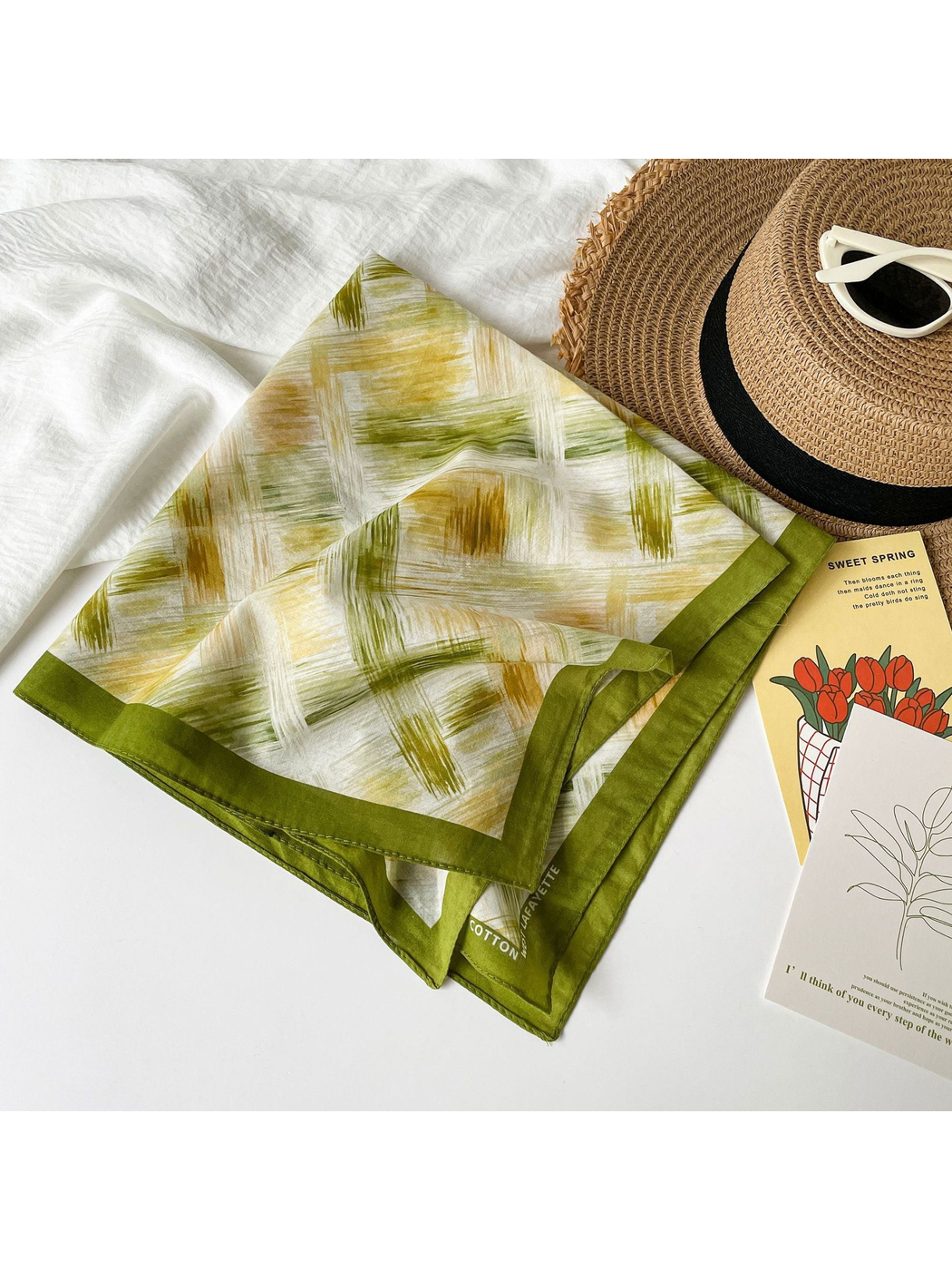 Olive green patterned square scarf draped beside a woven sun hat and sunglasses
