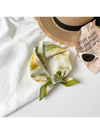 Olive green patterned square scarf tied in a loose knot beside a woven hat and sunglasses