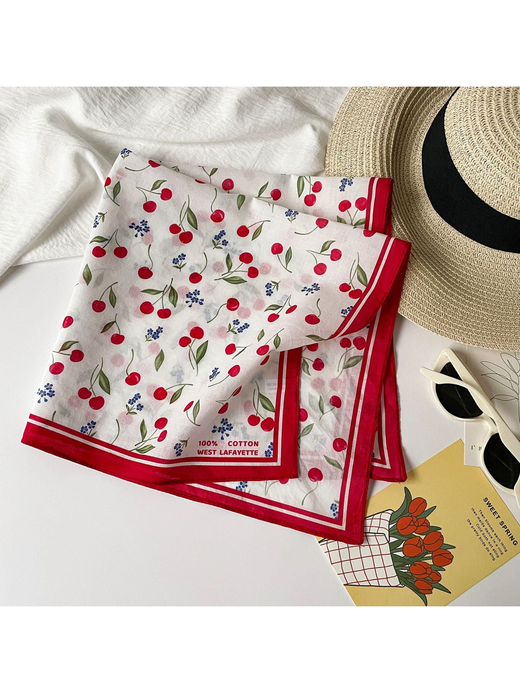 Cherry print square scarf folded beside a sun hat and sunglasses