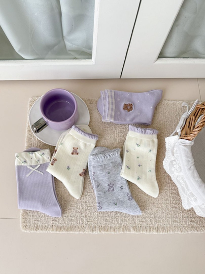 Five patterned crew sock styles in lavender, ivory, and heather gray arranged together