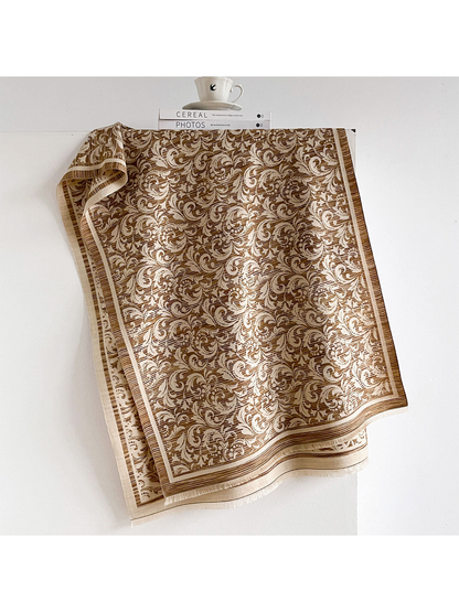 Brown scarf with cream scrolling leaves, a striped border and short fringe, draped open.