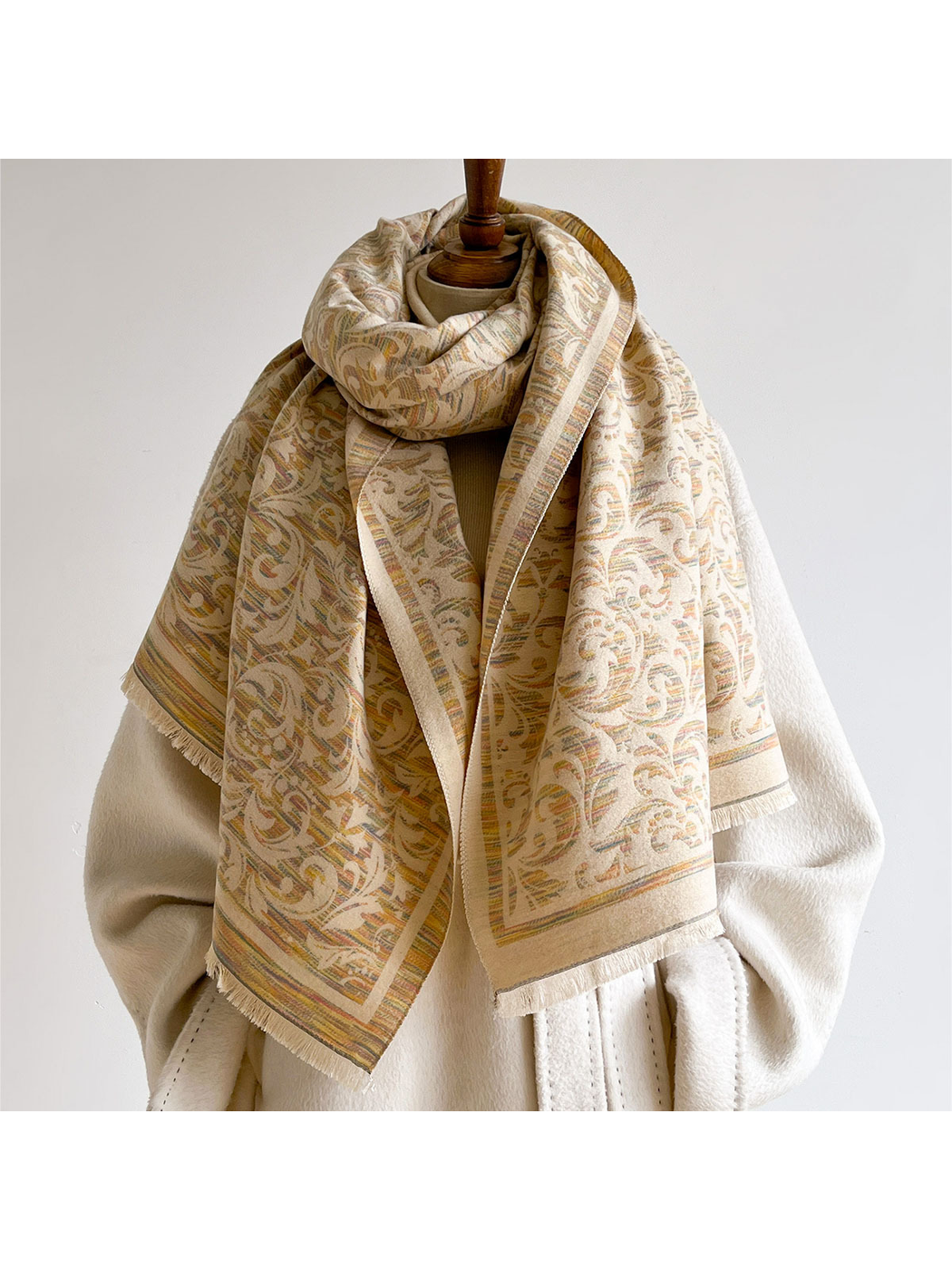 Khaki scrolling leaf scarf looped around a mannequin's neck with fringed ends draped over a cream coat.