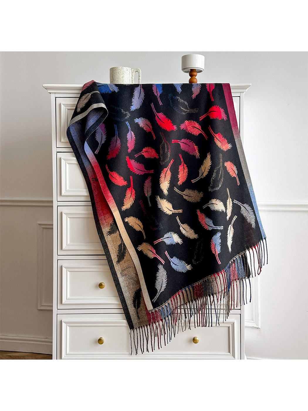 Black scarf with colorful feather motifs, contrasting borders and long fringe, draped over a white dresser.