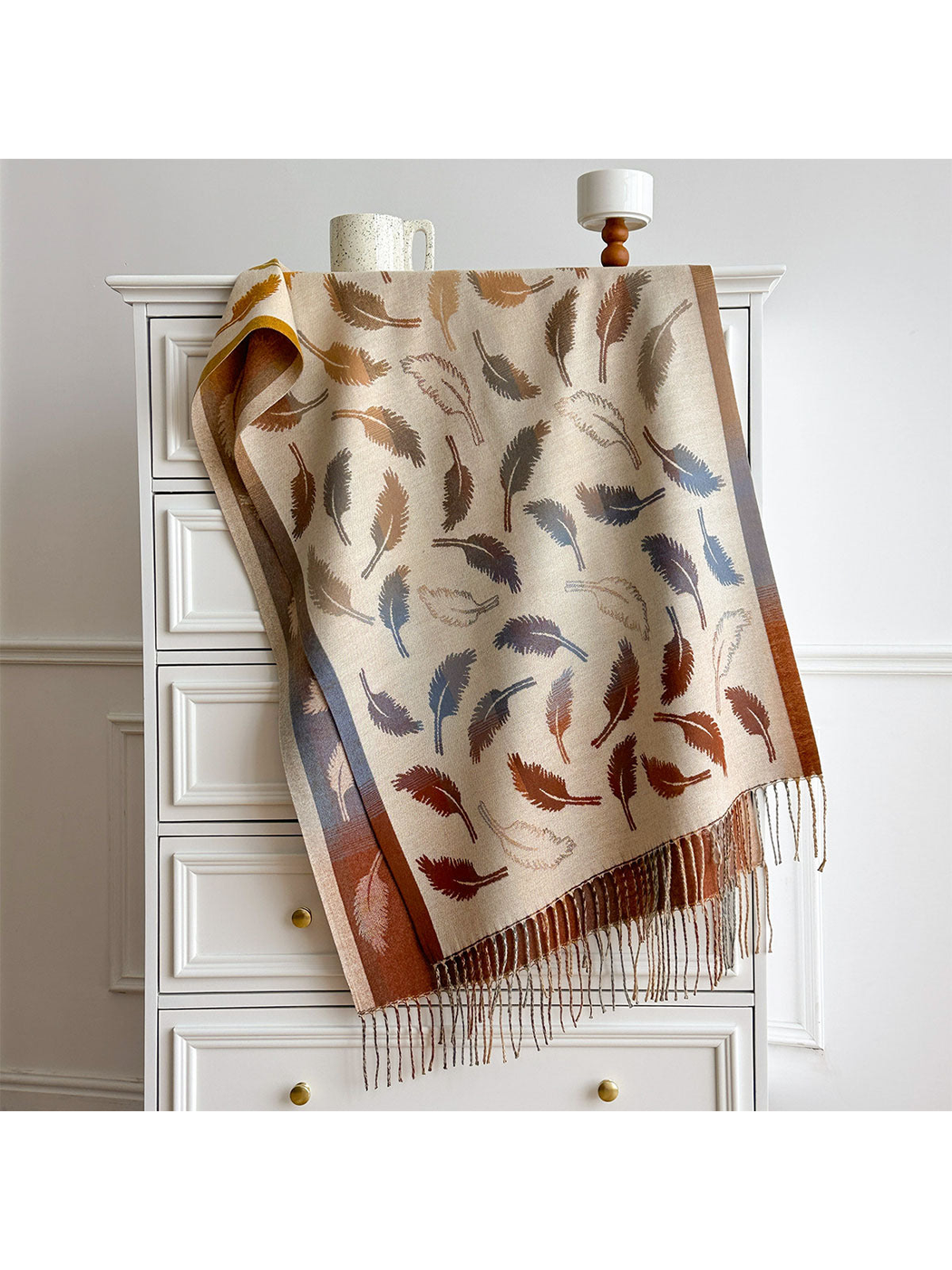 Cream feather-pattern scarf draped over a white dresser, with contrasting borders and twisted fringe.