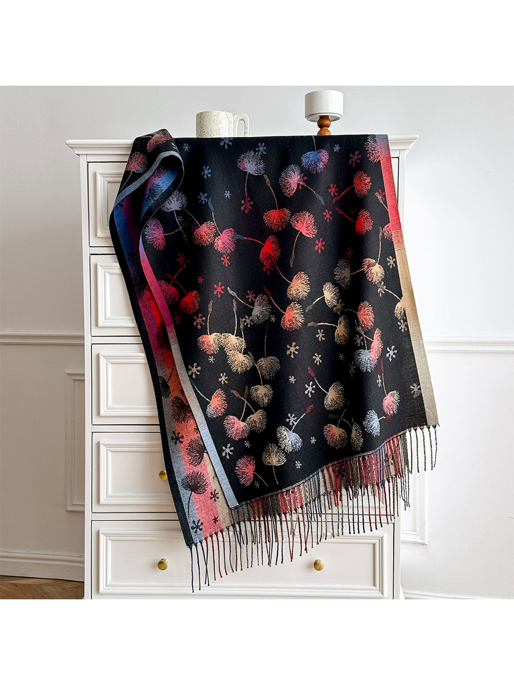 Black scarf with multicolored floral motifs, contrasting borders and long fringe, draped over a white chest.