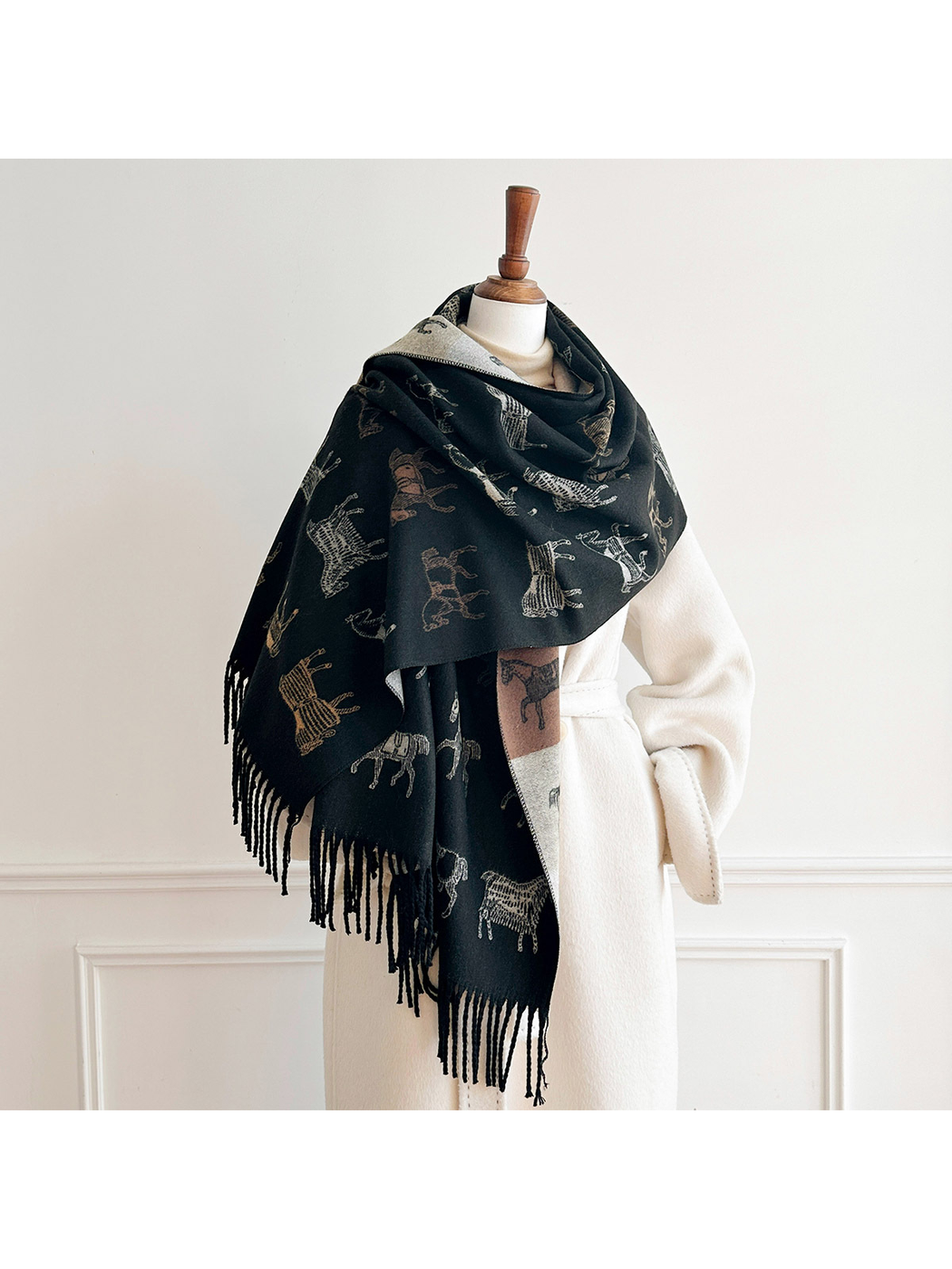 Black horse motif scarf draped broadly around a mannequin's shoulders over a cream coat, with fringed ends hanging down.