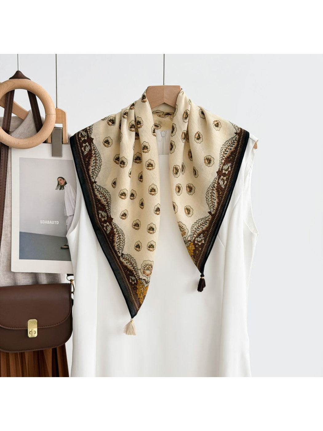 White floral scarf with an ornamental border and corner tassels draped over a white top on a hanger.