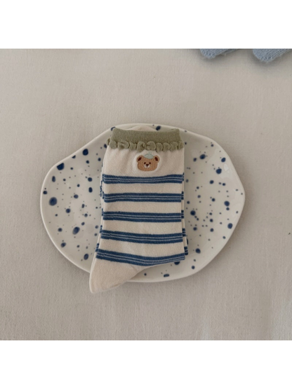Ivory socks with blue stripes, a bear motif and green scalloped cuffs, folded on a spotted plate.