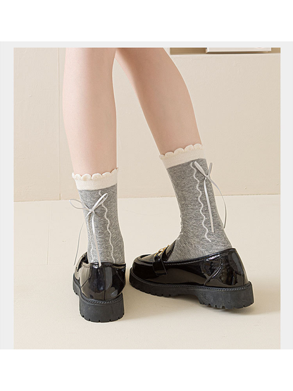 Gray wavy-pattern socks with ribbon bows and scalloped cuffs worn with glossy black loafers, rear view.