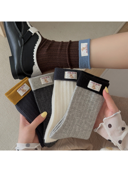 Ribbed socks in five colors with contrasting folded cuffs and bear labels, held beside a worn Dark Brown sock.