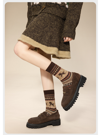 Coffee horse-pattern crew socks worn with brown loafers and a knit skirt, with one leg raised.