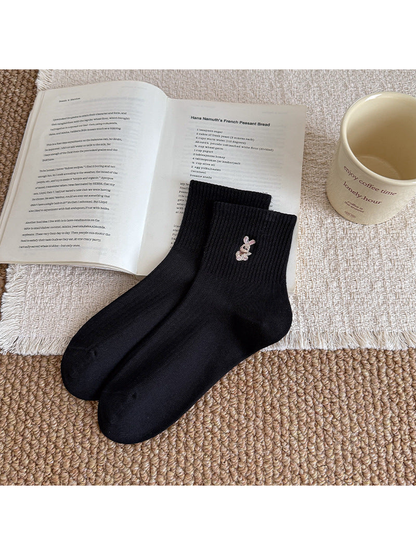 Black ribbed socks with seated-rabbit embroidery near the cuff, arranged beside an open book.