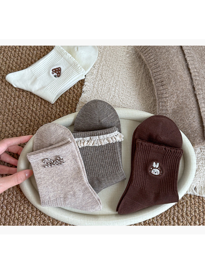 Folded Beige bow, Gray lace-trimmed and Dark Brown rabbit socks on a tray, with an Ivory mushroom-house sock nearby.