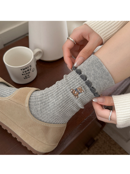 Close-up of a worn Gray sock as hands adjust its cuff, showing bear embroidery and dark loop trim.