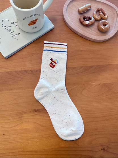Cup style ivory speckled sock with drink character embroidery and three colored cuff stripes, laid flat.