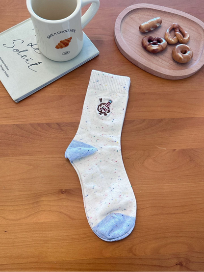 Dessert style ivory speckled sock with embroidered character and contrasting blue heel and toe, laid flat.