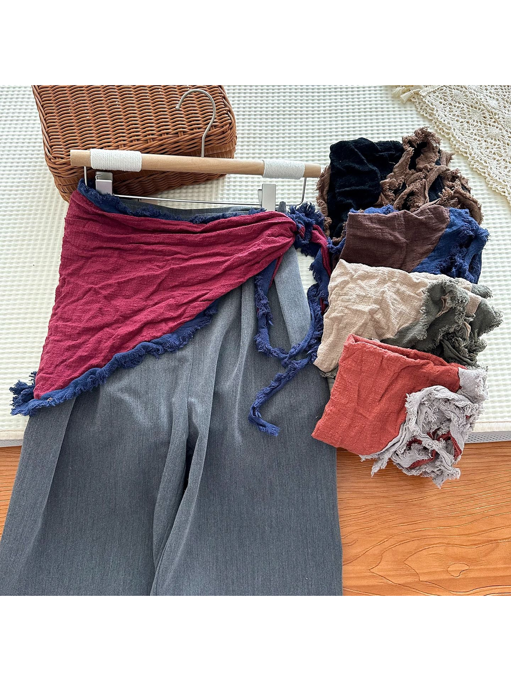 Burgundy scarf tied over gray pants beside folded scarves in contrasting colors.
