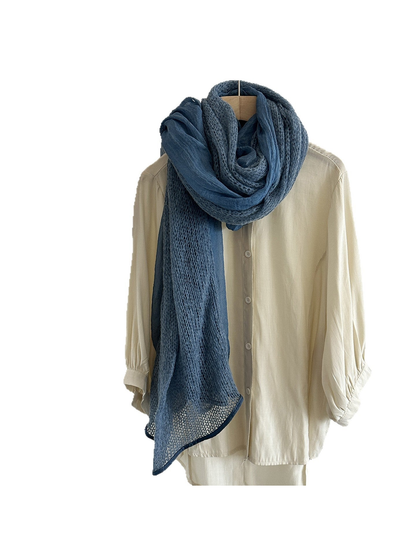 Blue scarf with openwork and sheer panels in a layered loop over a cream shirt against white.
