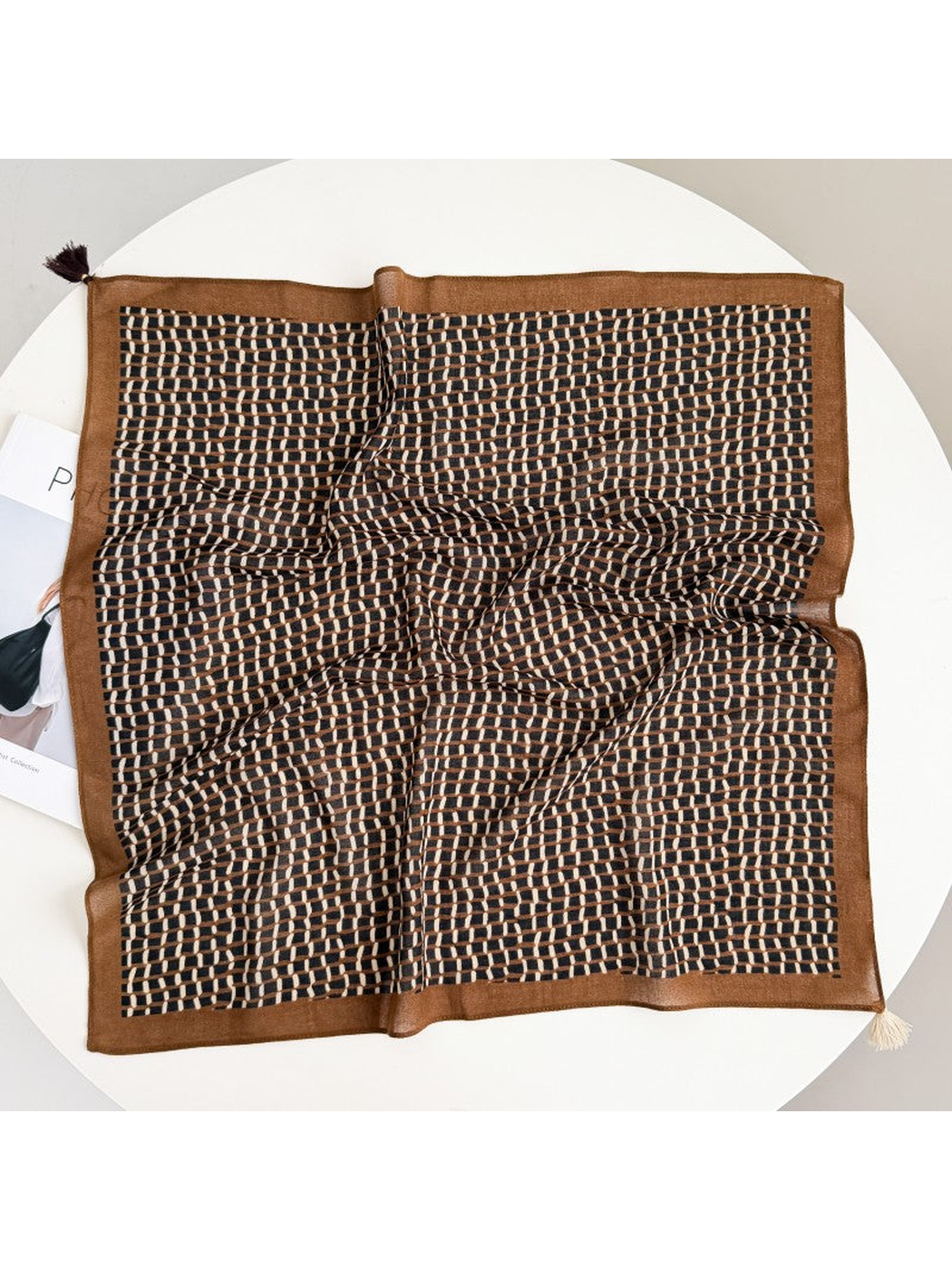 Brown square scarf with a black and cream geometric grid, wide border and two contrasting corner tassels, spread on a round table.