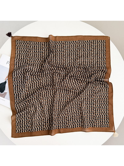 Brown square scarf with a black and cream geometric grid, wide border and two contrasting corner tassels, spread on a round table.