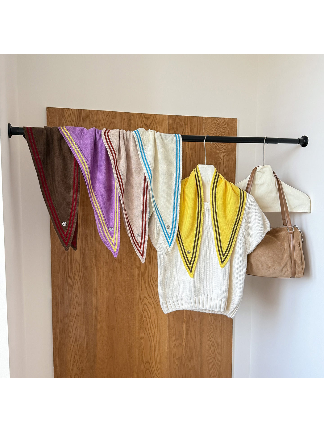 Five colors of striped-border triangle scarves hanging from a rail and draped over a white knit top.