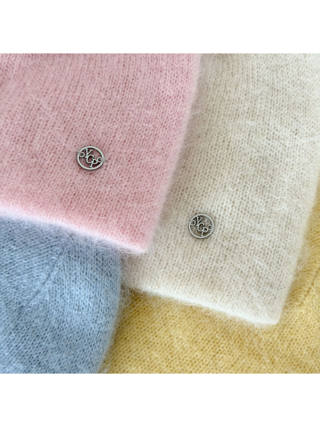 Close-up of fuzzy knit stitches, lower edges and round silver-tone emblems on pastel beanies.