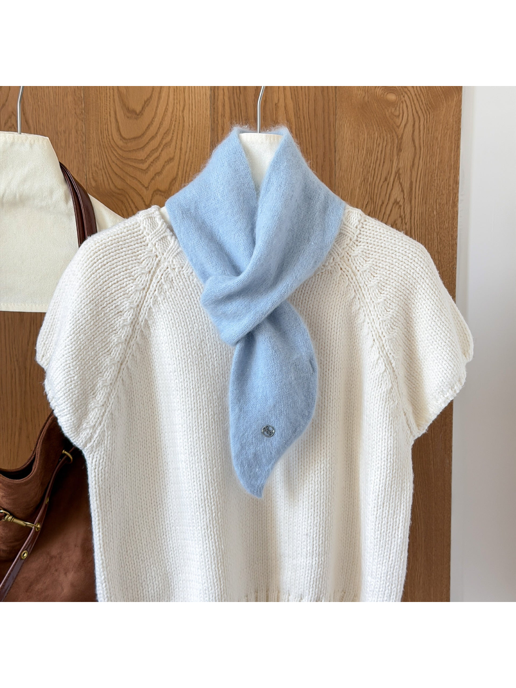 Sky Blue fuzzy knit scarf looped over a white knit top, with a tapered end and round silver-tone emblem.