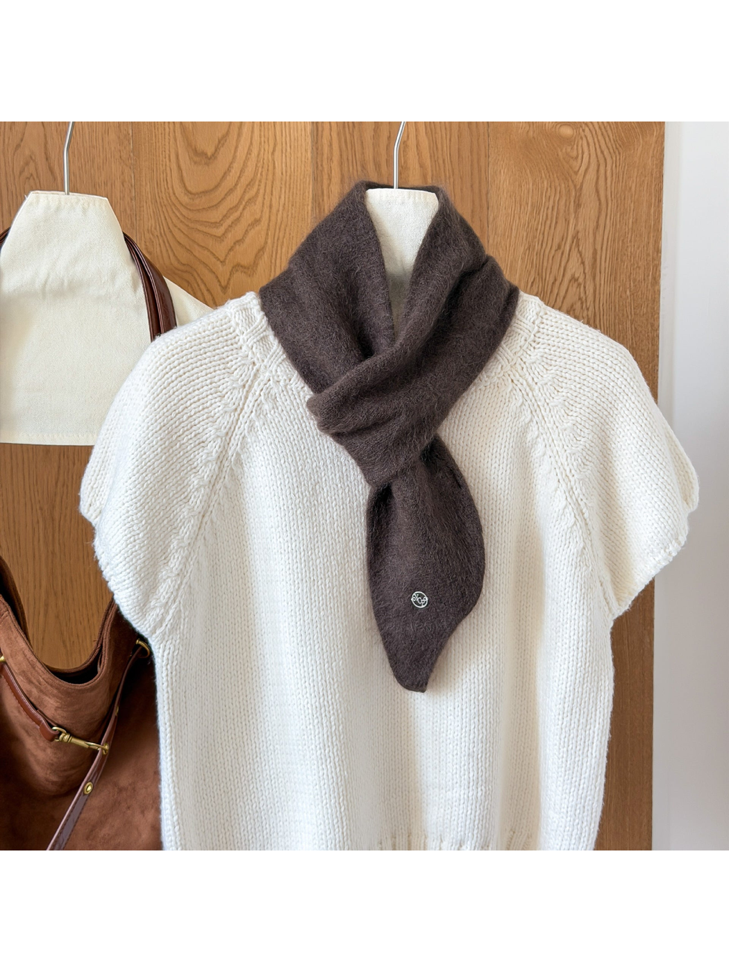 Dark Coffee fuzzy knit scarf looped over a white knit top, with a tapered end and round silver-tone emblem.